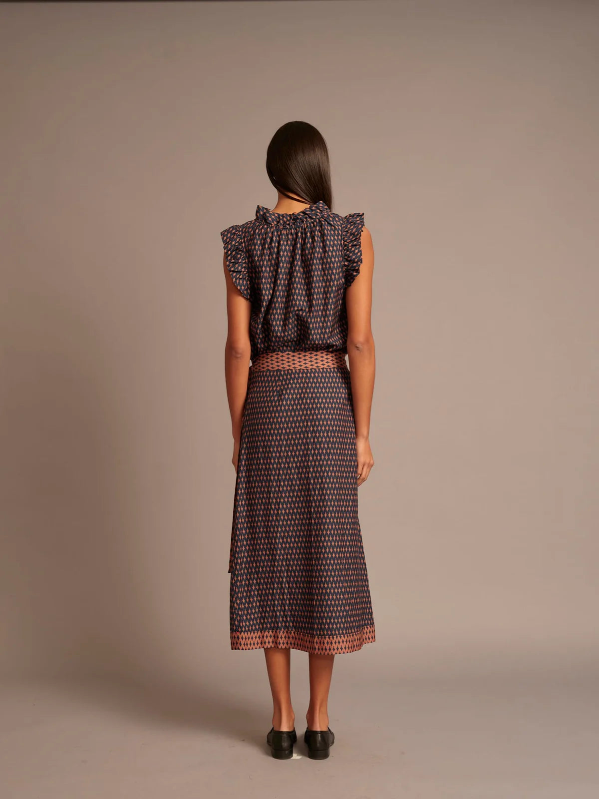 A woman viewed from the back wearing a sleeveless ruffled top and midi skirt set with a small-scale geometric pattern in dark purple and orange, set against a plain taupe studio background.