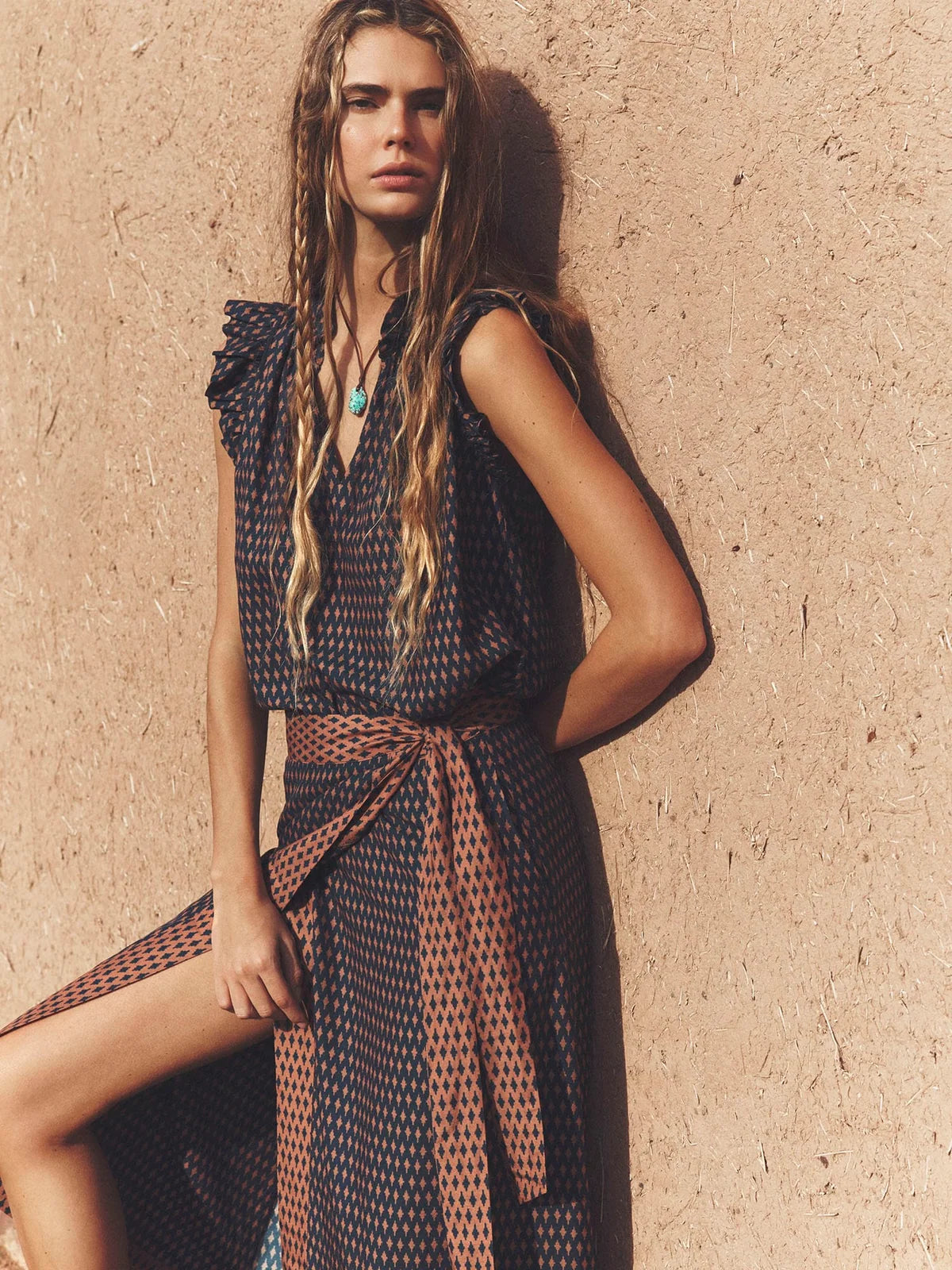 A woman models a sleeveless wrap-style midi dress with a dark navy and beige geometric pattern, featuring ruffled shoulders and a matching fabric tie belt at the waist, posed against a textured beige stucco wall.