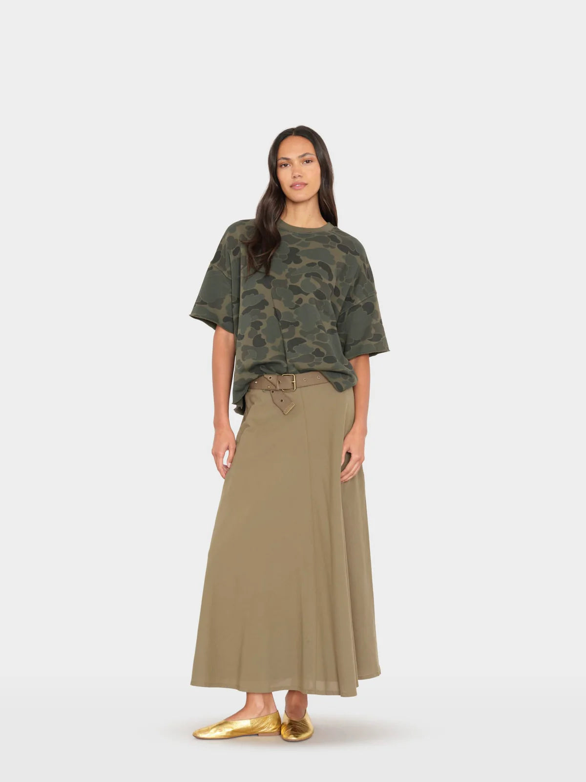 A woman modeling a short-sleeve camouflage t-shirt and a long, pleated olive green skirt, paired with metallic gold flats, against a light gray studio background.