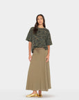 A woman modeling a short-sleeve camouflage t-shirt and a long, pleated olive green skirt, paired with metallic gold flats, against a light gray studio background.