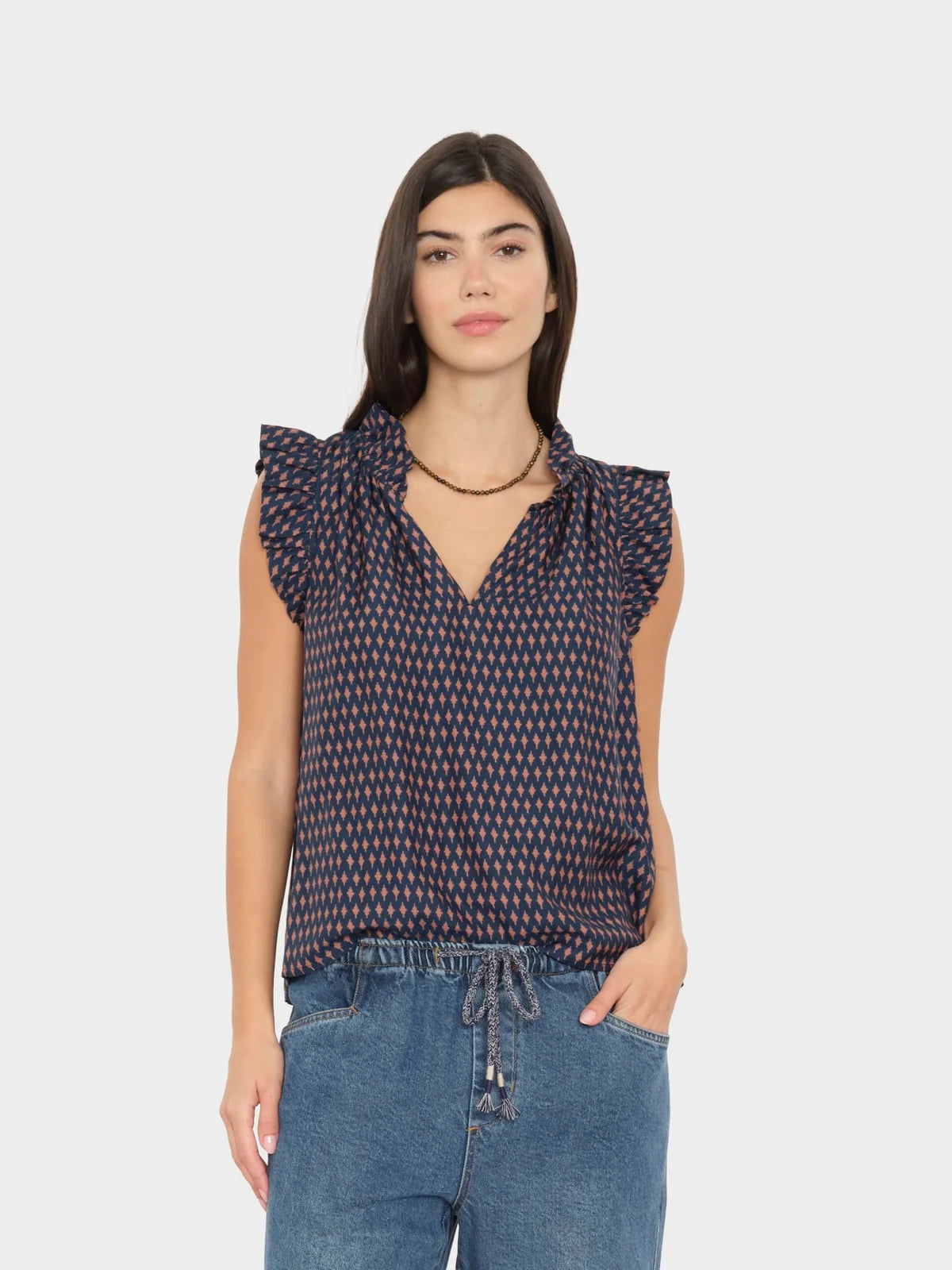 A woman modeling a sleeveless navy top with a small red and white geometric pattern, ruffled shoulders, and a V-neckline, paired with blue jeans against a light gray background.