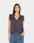 A woman modeling a sleeveless navy top with a small red and white geometric pattern, ruffled shoulders, and a V-neckline, paired with blue jeans against a light gray background.