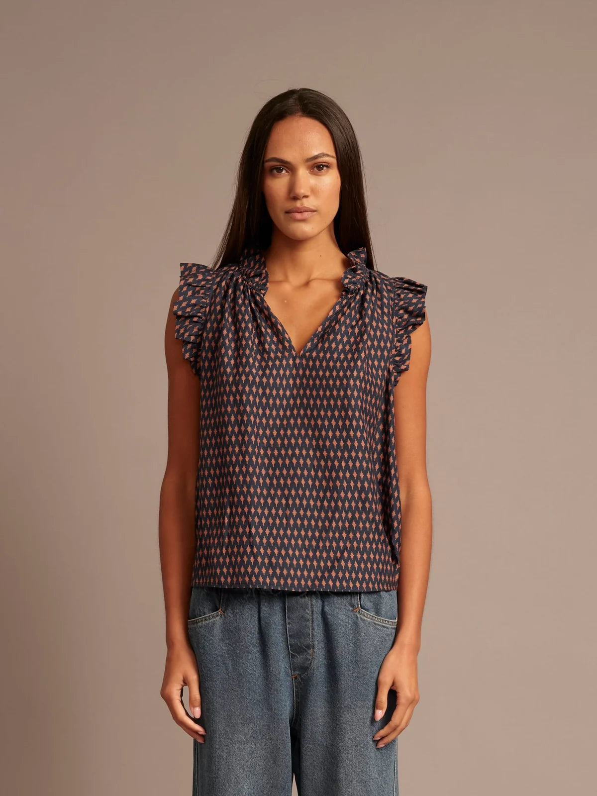 A woman modeling a sleeveless top with a small-scale geometric pattern in dark navy and red, featuring ruffled cap sleeves and a gathered V-neckline, paired with blue jeans against a solid taupe background.