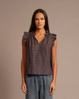 A woman modeling a sleeveless top with a small-scale geometric pattern in dark navy and red, featuring ruffled cap sleeves and a gathered V-neckline, paired with blue jeans against a solid taupe background.