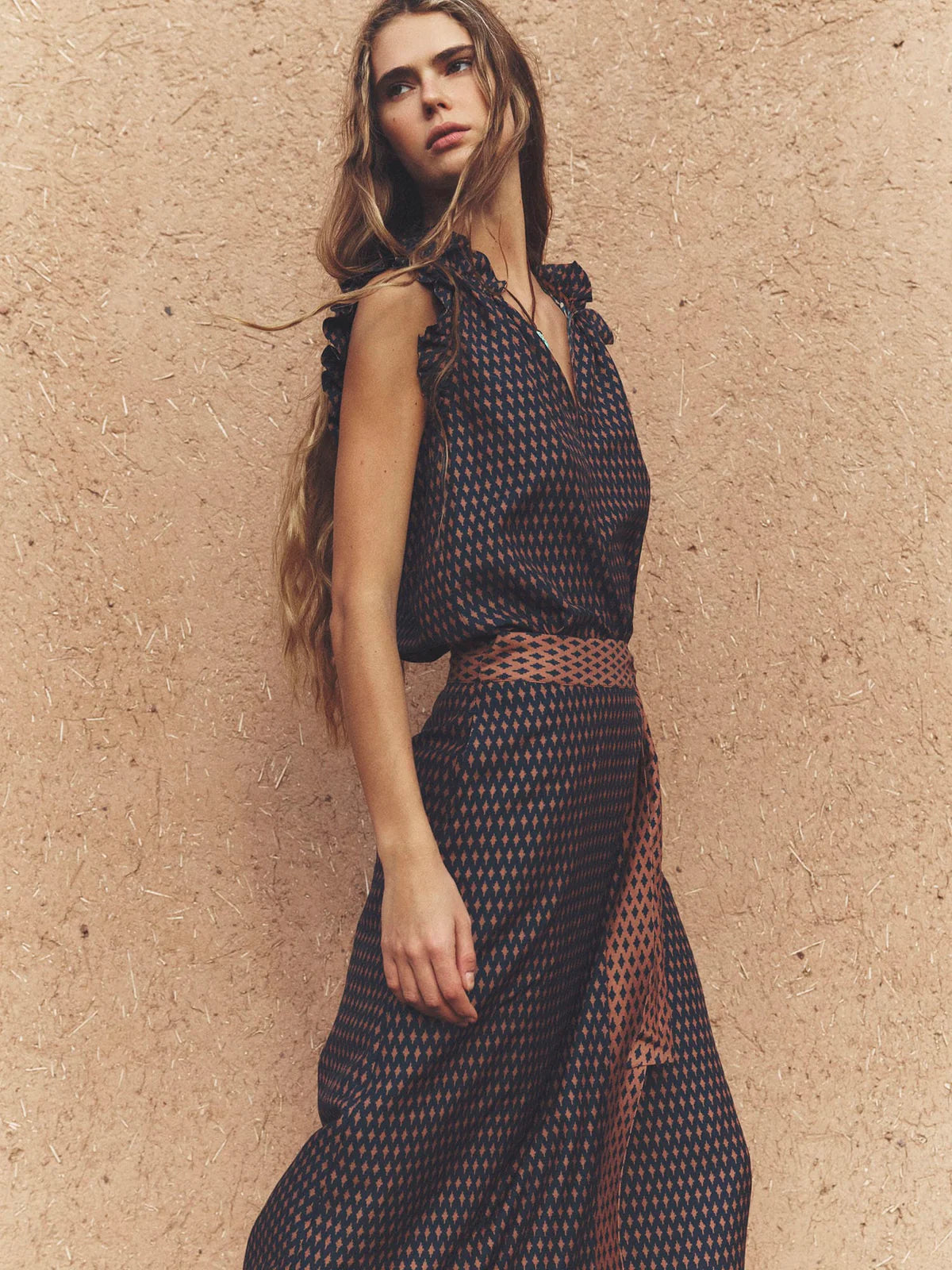 A woman wearing a sleeveless dark patterned dress with a sheer mesh belt, posing against a textured beige stucco wall.