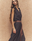 A woman wearing a sleeveless dark patterned dress with a sheer mesh belt, posing against a textured beige stucco wall.