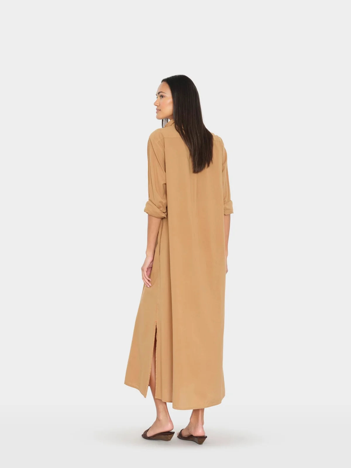A woman modeling a long, loose-fitting tan maxi dress from the back, featuring three-quarter sleeves rolled up to the elbow and a side slit, set against a plain light gray studio background.