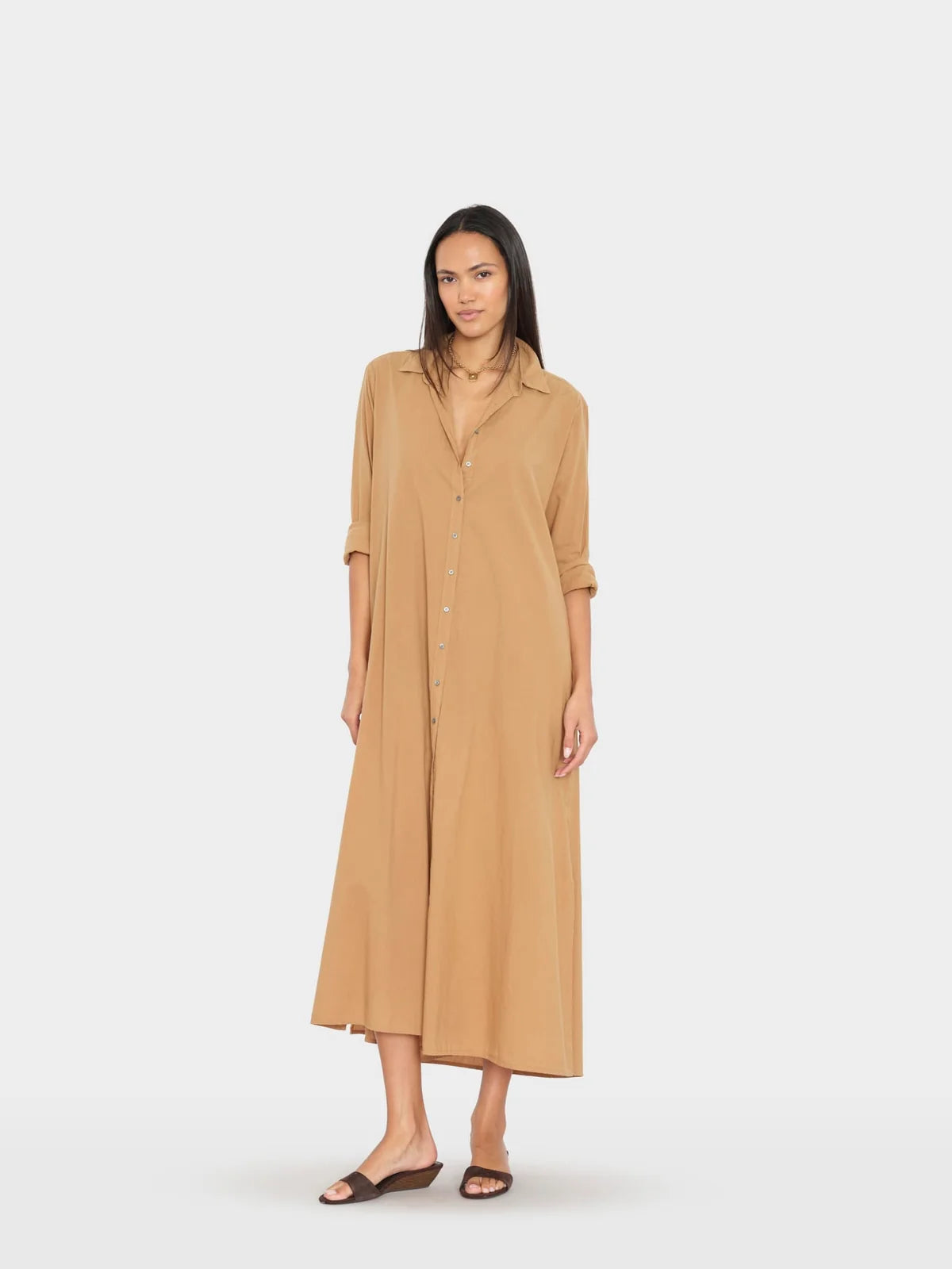A woman modeling a long tan button-front shirt dress with three-quarter sleeves and a relaxed midi-to-mud length, standing against a plain light gray studio background.
