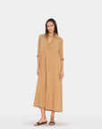 A woman modeling a long tan button-front shirt dress with three-quarter sleeves and a relaxed midi-to-mud length, standing against a plain light gray studio background.