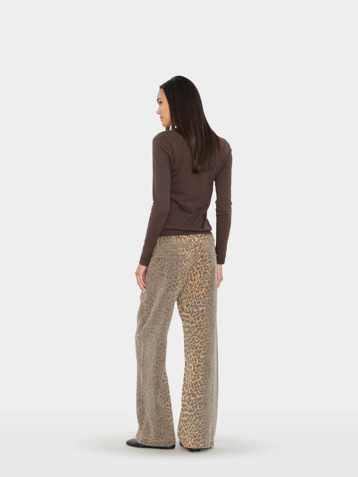 A woman is shown from the back wearing a dark brown long-sleeve top and beige wide-leg trousers with a leopard print pattern, set against a plain light gray studio background.