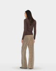 A woman is shown from the back wearing a dark brown long-sleeve top and beige wide-leg trousers with a leopard print pattern, set against a plain light gray studio background.