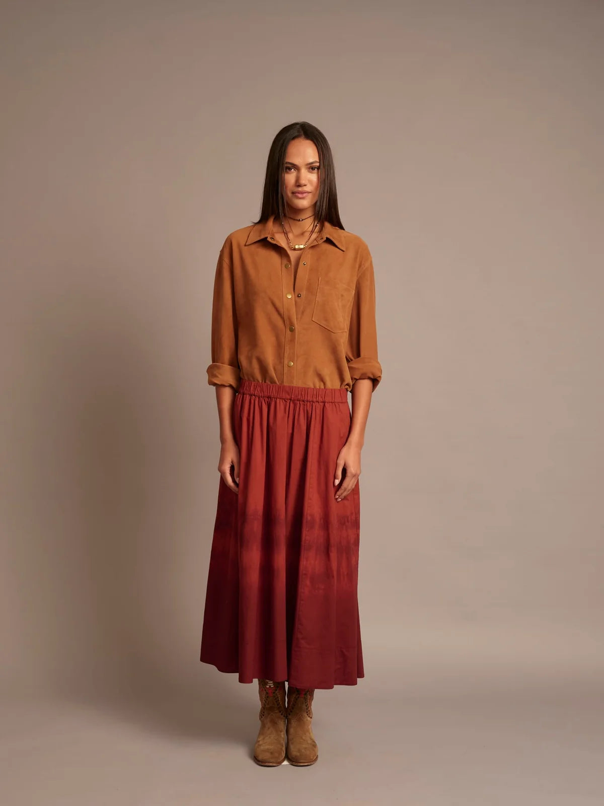 A woman stands against a plain warm beige studio background wearing a rust-brown button-up shirt and a long red skirt with a darker red gradient near the hem, paired with tan ankle boots.