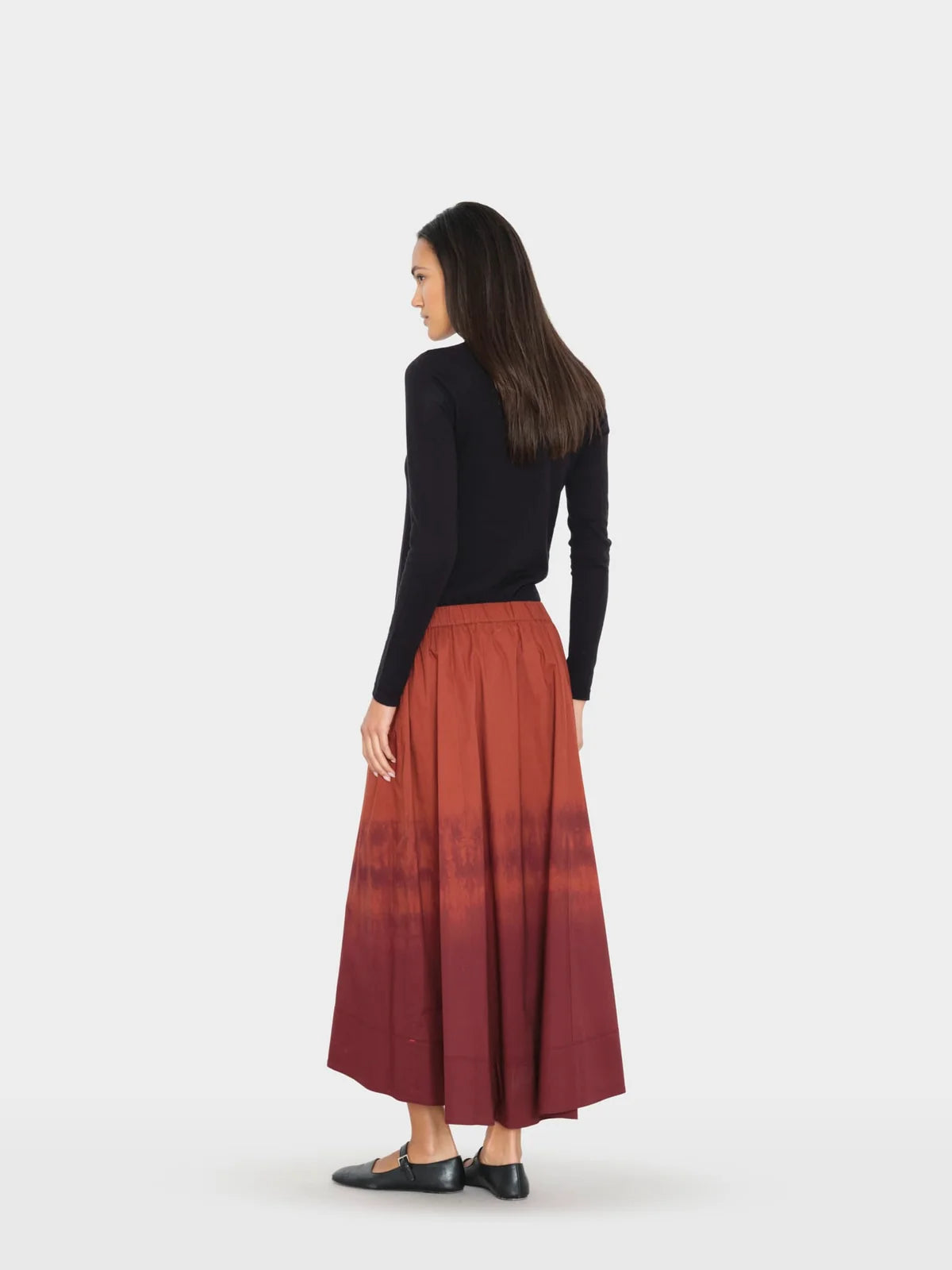A woman shown from the back wearing a black long-sleeve top and a long pleated skirt with a rust-orange to deep red gradient, standing against a light gray studio background.