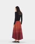 A woman shown from the back wearing a black long-sleeve top and a long pleated skirt with a rust-orange to deep red gradient, standing against a light gray studio background.