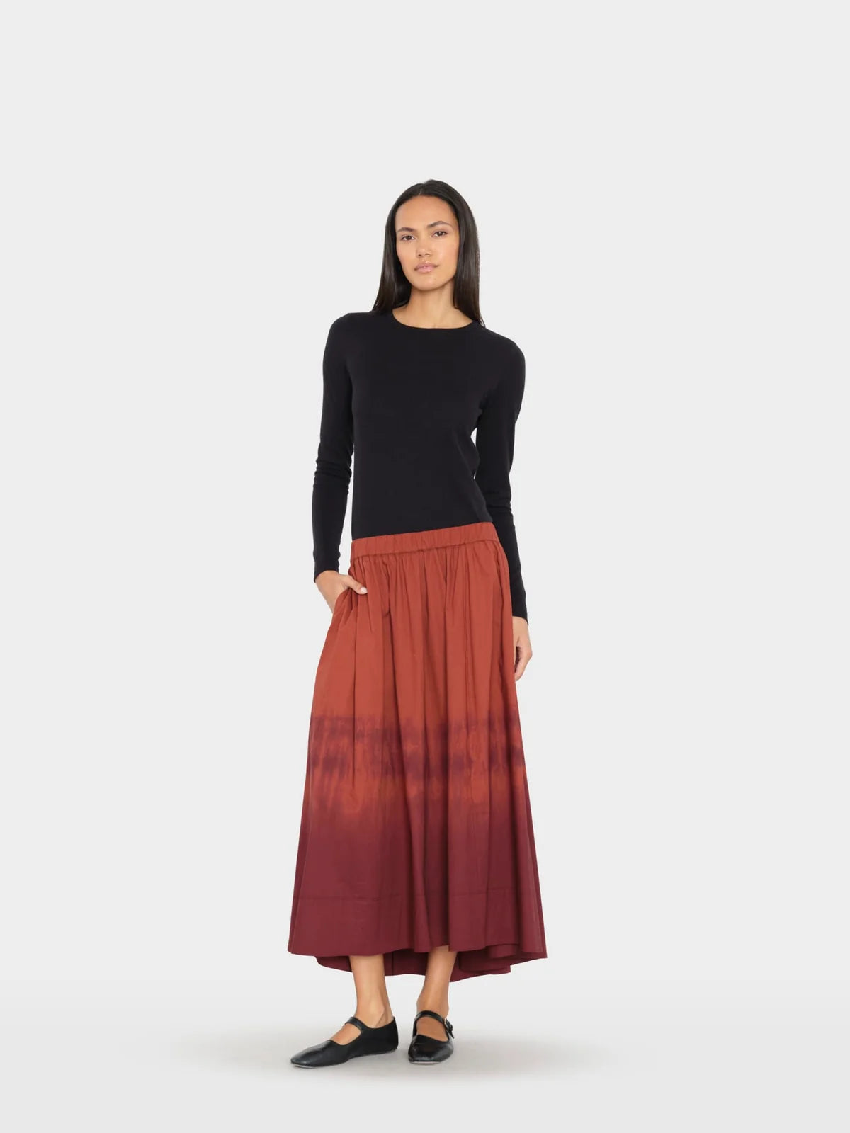 A woman modeling a long-sleeve black knit top and a pleated midi skirt in a rust-red to dark burgundy ombré tie-dye pattern, paired with black flats, against a light gray studio background.