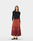 A woman modeling a long-sleeve black knit top and a pleated midi skirt in a rust-red to dark burgundy ombré tie-dye pattern, paired with black flats, against a light gray studio background.
