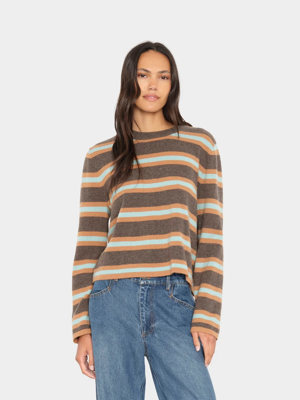 A woman modeling a long-sleeve knit sweater with horizontal stripes in brown, beige, and light blue, paired with blue jeans, against a light gray studio background.