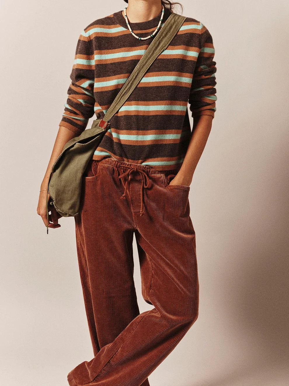 A person wearing a brown, beige, and white striped long-sleeve knit top, rust-brown corduroy drawstring pants, and an olive green crossbody bag, photographed against a plain beige studio background.