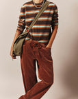 A person wearing a brown, beige, and white striped long-sleeve knit top, rust-brown corduroy drawstring pants, and an olive green crossbody bag, photographed against a plain beige studio background.
