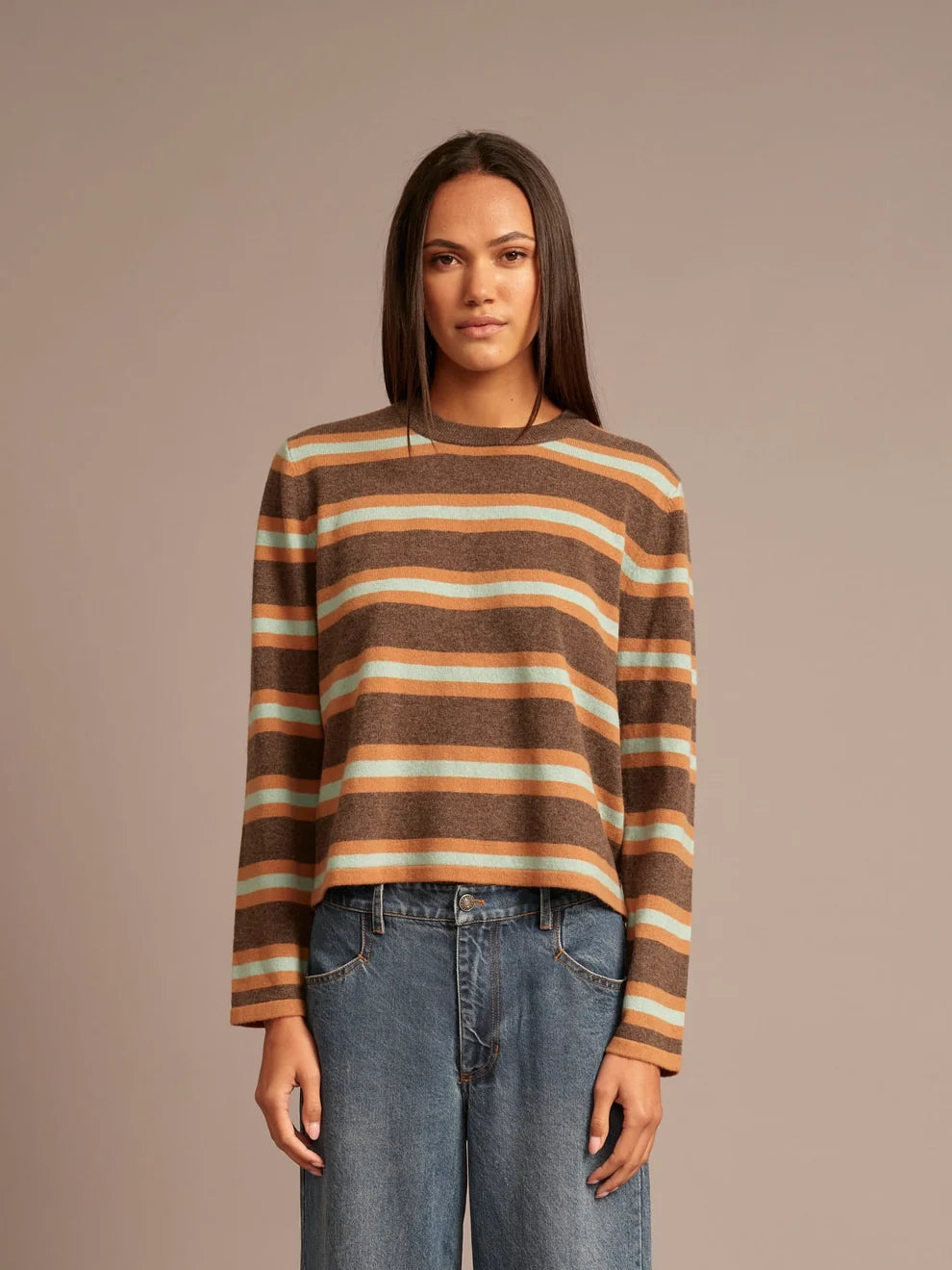 A woman modeling a cropped, long-sleeve knit sweater with horizontal stripes in brown, beige, and orange, paired with blue jeans, against a solid taupe background.
