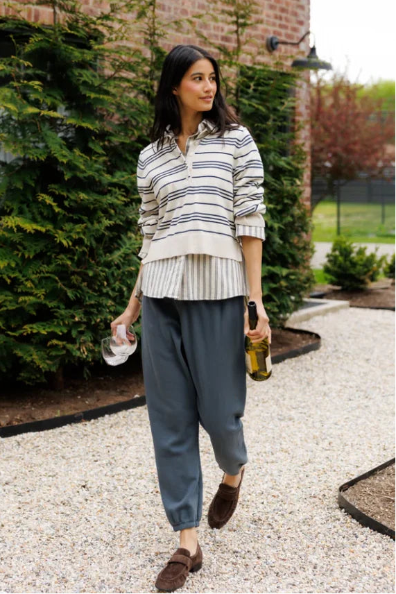 A woman walking on a gravel path in front of a brick house, wearing a cream and navy striped long-sleeve button-up shirt, loose dark gray pants, and brown loafers.