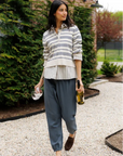 A woman walking on a gravel path in front of a brick house, wearing a cream and navy striped long-sleeve button-up shirt, loose dark gray pants, and brown loafers.