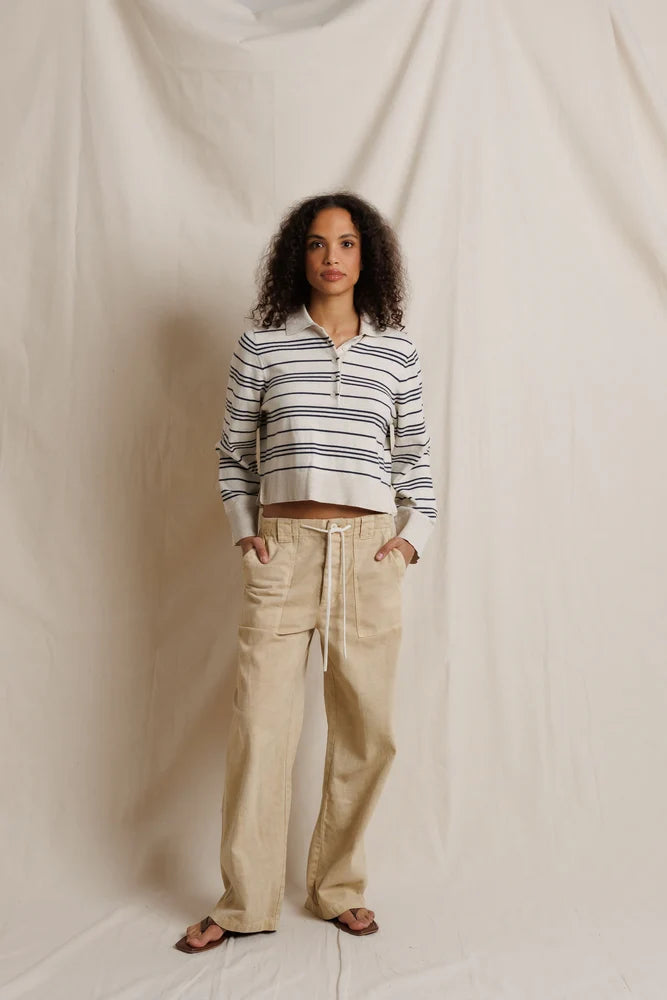 A woman stands against a draped cream fabric backdrop wearing a cropped long-sleeve polo-style top with navy and beige horizontal stripes, paired with loose beige pants and dark sandals.