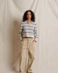 A woman stands against a draped cream fabric backdrop wearing a cropped long-sleeve polo-style top with navy and beige horizontal stripes, paired with loose beige pants and dark sandals.