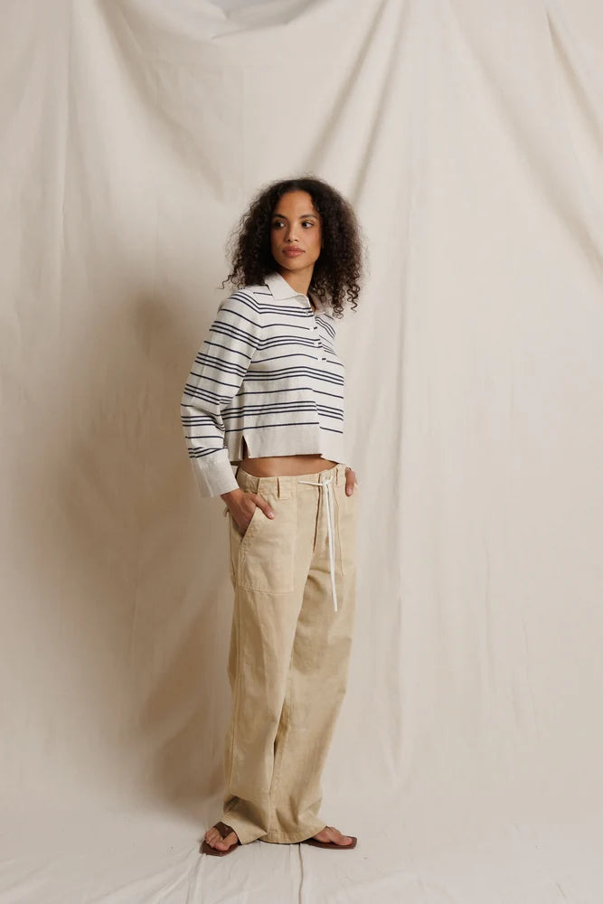 A woman stands against a draped cream fabric backdrop wearing a cropped long-sleeve striped knit top with navy and white stripes, paired with loose beige drawstring pants and brown sandals.