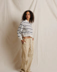 A woman stands against a draped cream fabric backdrop wearing a cropped long-sleeve striped knit top with navy and white stripes, paired with loose beige drawstring pants and brown sandals.