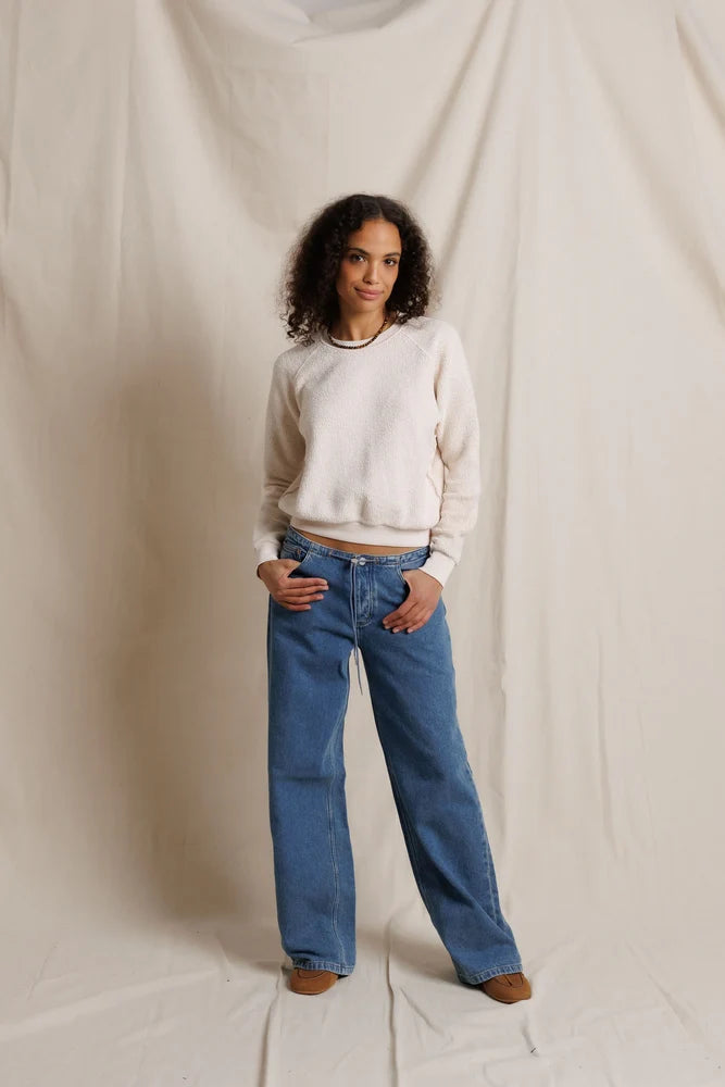A woman stands against a draped cream fabric backdrop wearing a cream crewneck sweater, blue wide-leg jeans, and brown shoes.
