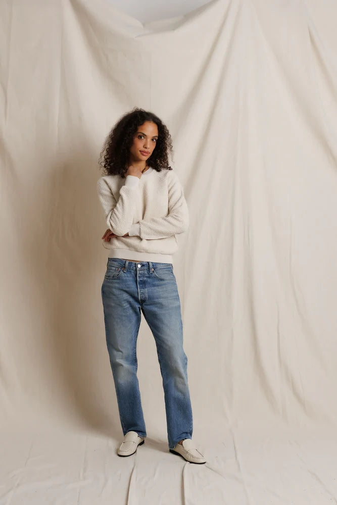 A woman with curly dark hair stands in front of a draped off-white fabric backdrop, wearing a cream long-sleeve knit top, blue straight-leg jeans, and off-white slip-on shoes.