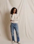 A woman with curly dark hair stands in front of a draped off-white fabric backdrop, wearing a cream long-sleeve knit top, blue straight-leg jeans, and off-white slip-on shoes.