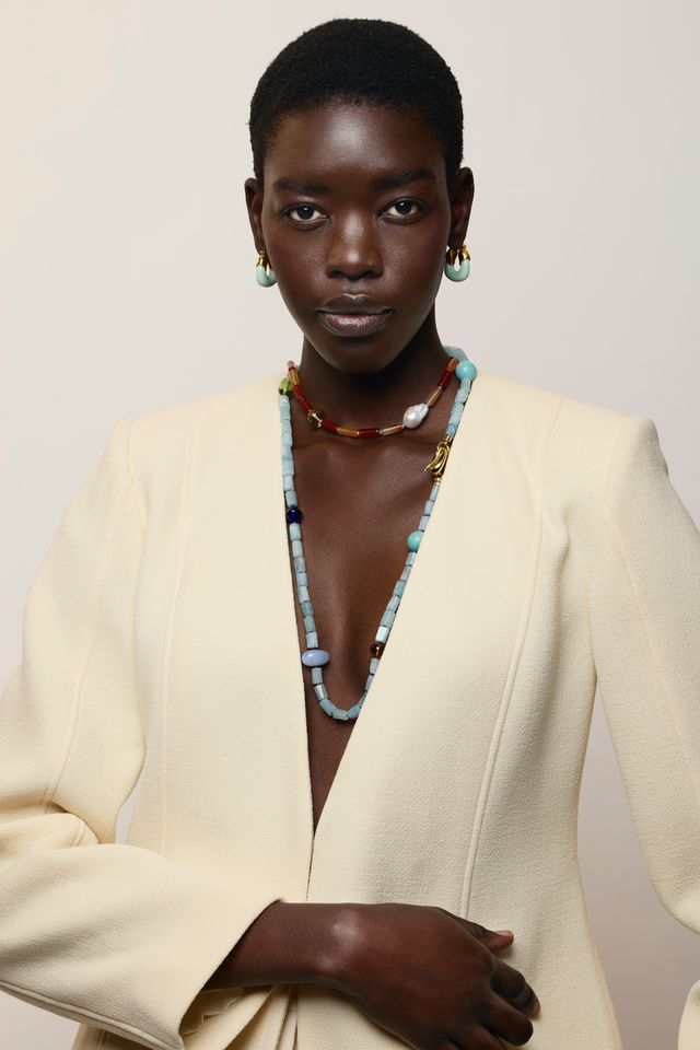 A model with short dark hair is wearing a cream-colored blazer, Mini Organic Hoops in Seafoam and layered beaded necklaces, including a prominent light blue beaded necklace, against a plain light beige background.